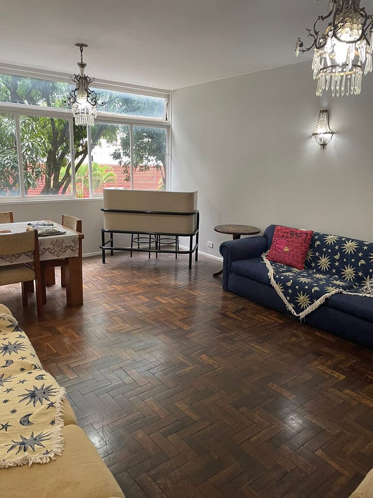 Uma sala de estar com piso de parquet, dois sofás com capas estampadas, uma mesa de jantar de madeira com cadeiras, um piano perto de uma grande janela e dois lustres pendurados no teto. Árvores são visíveis do lado de fora da janela.
