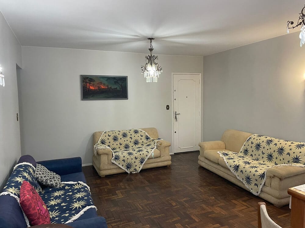 Uma sala de estar com dois sofás bege e uma poltrona azul, todos revestidos com mantas estampadas. Há uma porta branca, piso de parquet, quadros nas paredes e lustres pendurados no teto.