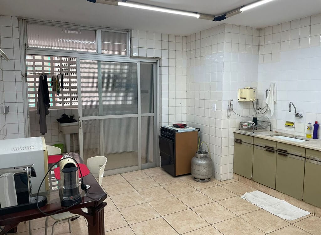 Uma cozinha com paredes e piso de azulejo, uma mesa com cadeiras, um micro-ondas, uma cafeteira, um fogão, um botijão de gás, uma pia, produtos de limpeza e roupas penduradas perto de portas de correr de vidro fosco.