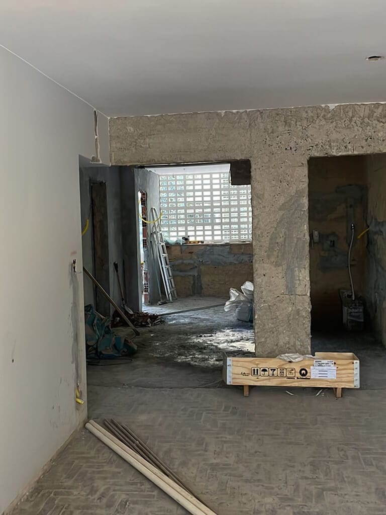 Uma sala parcialmente reformada com paredes de concreto expostas e materiais de construção espalhados, incluindo bolsas, baldes, ferramentas e uma pequena escada perto de uma janela com um padrão de grade.