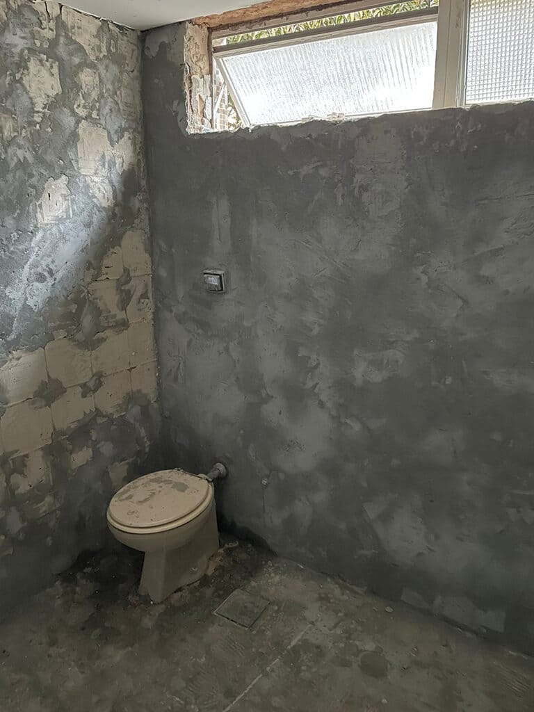 Um banheiro em reforma com paredes de concreto inacabadas, azulejos faltando, um vaso sanitário antigo, uma janela pequena e restos de construção no chão.