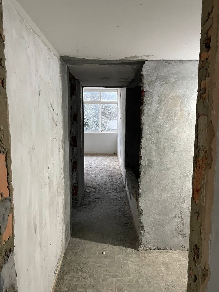 Um corredor estreito e inacabado, com paredes ásperas e parcialmente rebocadas, leva a uma janela que deixa entrar luz natural no fundo. O piso parece inacabado e os materiais de construção são visíveis.