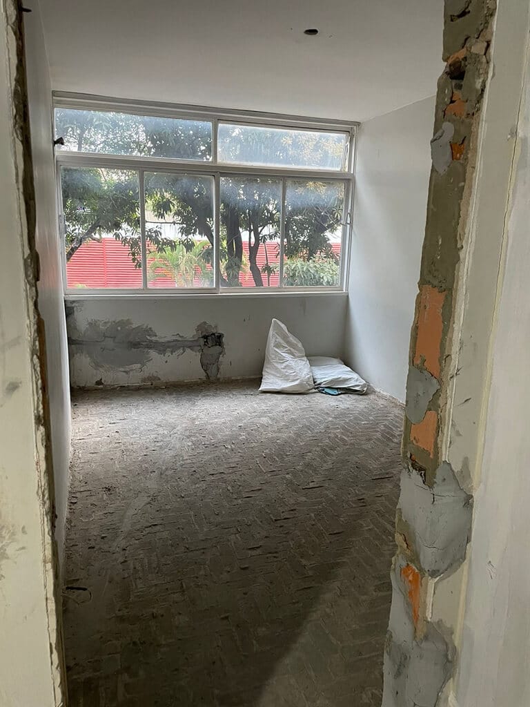 Um cômodo pequeno e inacabado, com piso de tijolos aparentes, paredes parcialmente rebocadas e janelas grandes. Duas almofadas ou sacolas brancas repousam no chão perto da janela, e árvores são visíveis do lado de fora.
