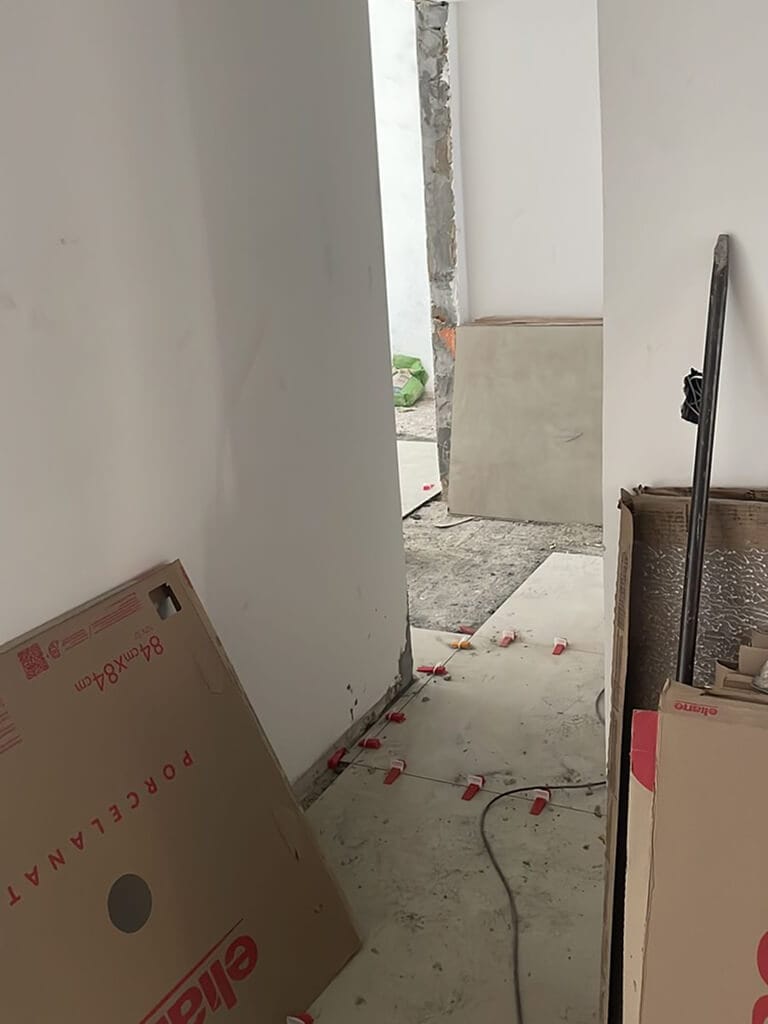 Um corredor em construção com azulejos soltos, caixas de papelão e ferramentas espalhadas pelo chão. As paredes são brancas e inacabadas, e uma porta aberta leva a outro cômodo.