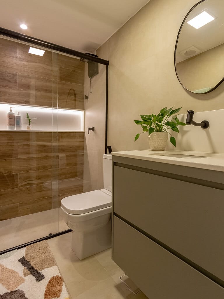 Banheiro moderno com box de vidro, azulejos que imitam madeira, pia suspensa com pia branca, espelho redondo, vaso sanitário branco e um vaso de plantas na bancada. No chão, um tapete estampado.
