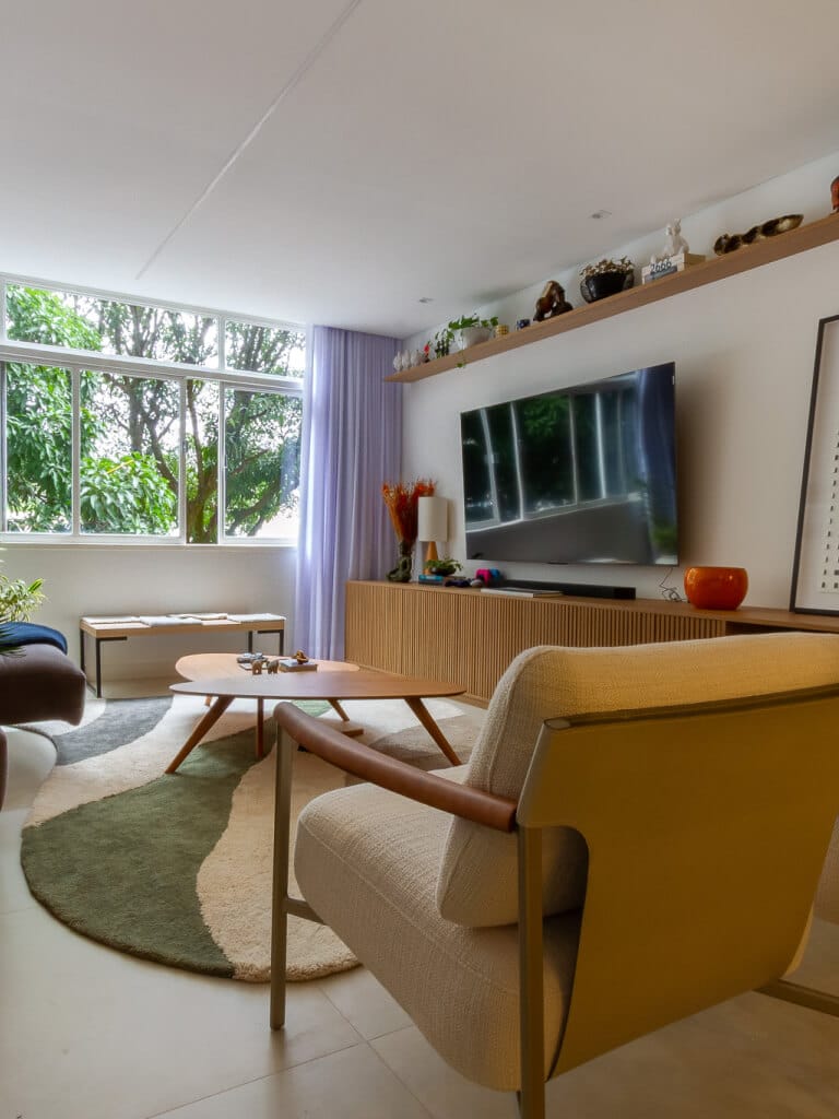 Sala de estar moderna com uma janela grande, TV de tela plana, móveis de cores claras, mesa redonda e tapete verde abstrato. Prateleiras com itens de decoração e árvores visíveis do lado de fora da janela adicionam uma sensação aconchegante e convidativa.