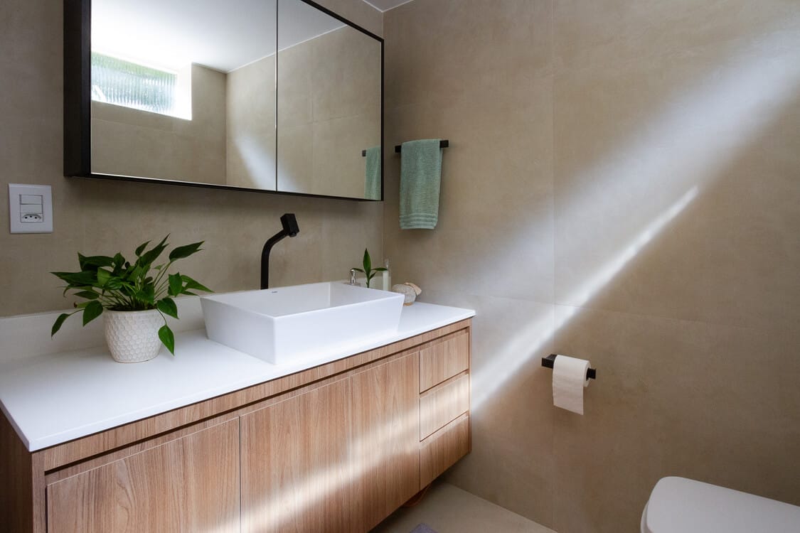 Banheiro moderno com paredes bege, uma pia de madeira com pia branca, uma torneira preta, um vaso de planta, um armário espelhado, um toalheiro com uma toalha verde e luz do sol entrando, projetando uma sombra diagonal brilhante.