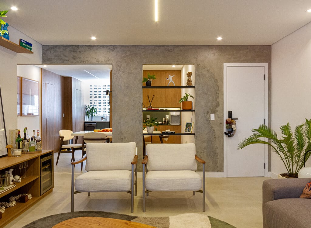 Sala de estar moderna com duas poltronas brancas, uma prateleira embutida em uma parede cinza, uma porta branca e uma planta. O ambiente se abre para uma sala de jantar e cozinha ao fundo, com iluminação aconchegante por toda parte.