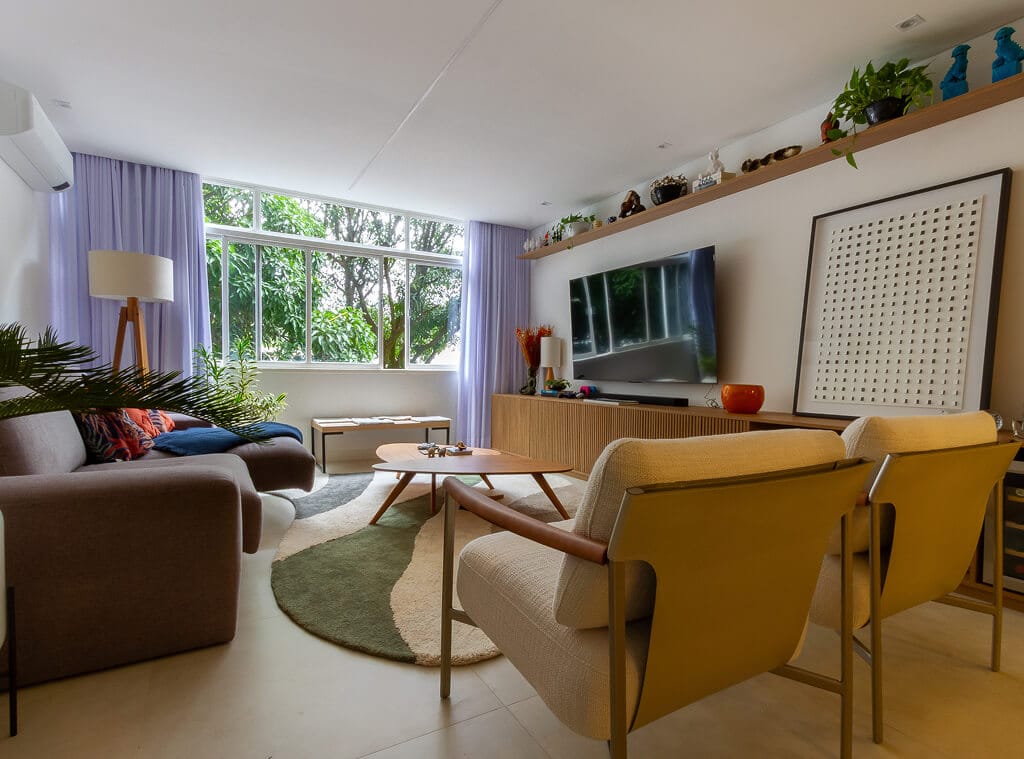 Sala de estar moderna com janela ampla, duas poltronas amarelas, um sofá marrom, um tapete redondo, uma mesa de centro de madeira, TV sobre um console, plantas e itens decorativos em uma prateleira e mesa lateral. A luz natural preenche o espaço.