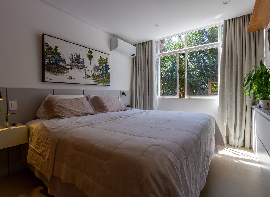 Uma cama impecavelmente arrumada com roupa de cama clara repousa em um quarto iluminado. Grandes janelas com cortinas deixam entrar a luz natural e revelam árvores verdes lá fora. Uma pintura está pendurada acima da cama e uma planta está sobre uma cômoda à direita.