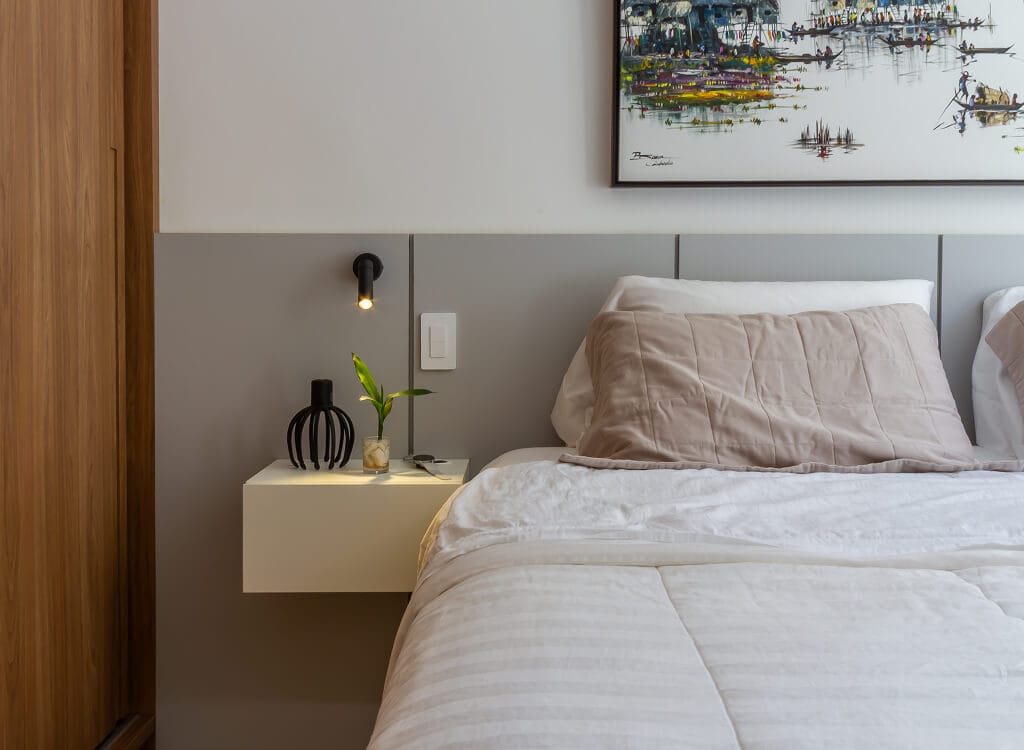 Um quarto moderno com uma cama bem arrumada, travesseiros bege, cabeceira cinza, mesa de cabeceira branca flutuante com uma pequena planta, vaso preto e luminária de parede; uma obra de arte emoldurada está pendurada acima.