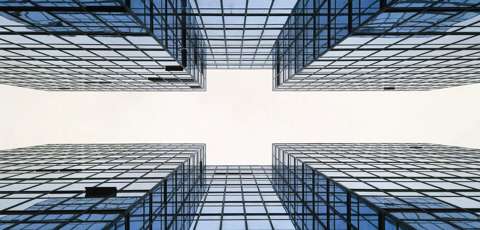 Uma vista de ângulo baixo de dois arranha-céus altos e modernos com fachadas de vidro. Os edifícios formam um V simétrico contra o céu branco, criando uma perspectiva arquitetônica impressionante. As janelas refletem a luz, adicionando um tom azul às estruturas.